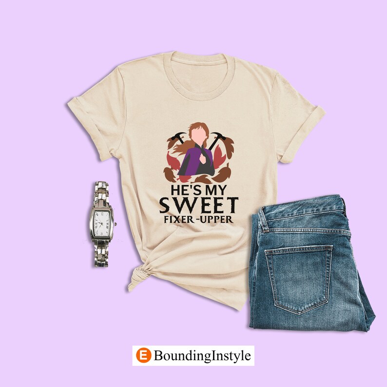 May include: A beige t-shirt with a graphic of a cartoon character with brown hair and a purple dress. The text "He's my sweet fixer-upper" is printed below the graphic.