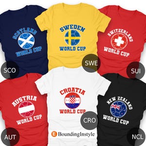 May include: A collection of six t-shirts, each featuring a soccer ball design with a country's name and "World Cup" text. Colors include navy, yellow, red, white, and black. The shirts display the flags of Scotland, Sweden, Switzerland, Austria, Croatia, and New Zealand.