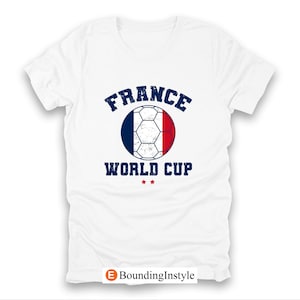 May include: White t-shirt featuring a soccer ball design in the colors of the French flag. The text "FRANCE WORLD CUP" is printed above and below the ball in a distressed navy blue font. Two small red stars are below the text.
