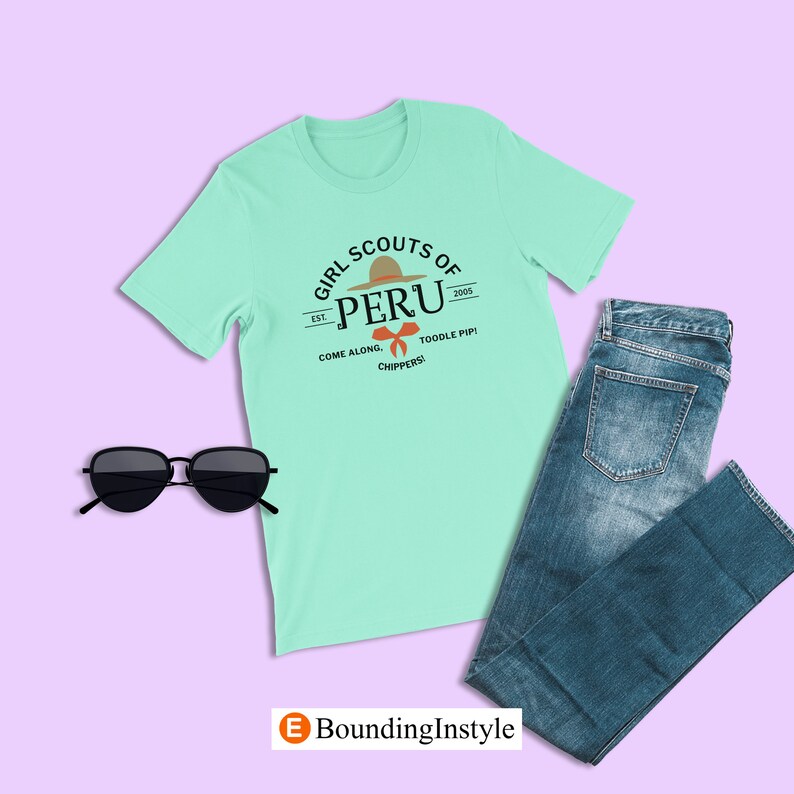 May include: A light green t-shirt featuring a graphic design of a Girl Scouts of Peru logo. The logo depicts a brown hat with an orange band and a red bow. The text on the logo reads "Girl Scouts of Peru Est. 2005 Come Along. Toodle Pip! Chippers!"