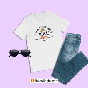 May include: White t-shirt with a graphic design of a Girl Scout patch. The patch features a brown hat with a red bow and the text "Girl Scouts of Peru" with the text "Est. 2005" and "Come Along, Toodle Pip! Chippers!" underneath.