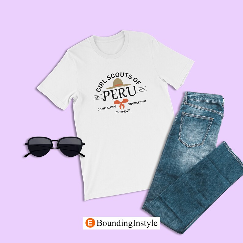 May include: White t-shirt with a graphic design of a Girl Scout patch. The patch features a brown hat with a red bow and the text "Girl Scouts of Peru" with the text "Est. 2005" and "Come Along, Toodle Pip! Chippers!" underneath.