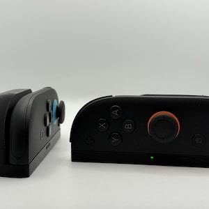 May include: Two black game controllers, one with buttons and an orange joystick, the other with a blue joystick. The controllers are on a white surface. The controllers are for a gaming console.