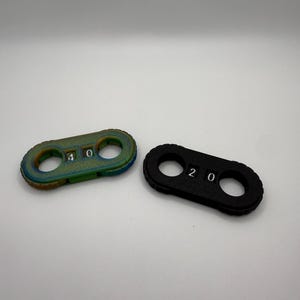 May include: Two small, oblong-shaped objects. One is a mix of green, blue, and gold with the numbers "4 0" printed on it. The other is black with the numbers "2 0" printed on it. Both have two circular holes.