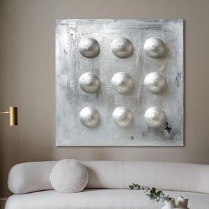 May include: A contemporary living room with a white sofa, glass coffee table, and abstract artwork. The artwork is a square canvas with a textured silver background and nine raised, spherical silver elements. A gold floor lamp and patterned rug complete the stylish interior.