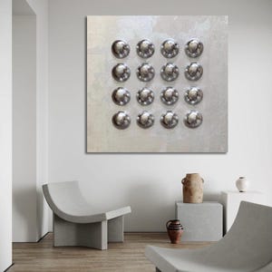 May include: A square, textured canvas features a grid of sixteen shiny, silver, dome-shaped objects. The artwork is displayed in a minimalist room with two curved, gray concrete chairs and decorative pottery.