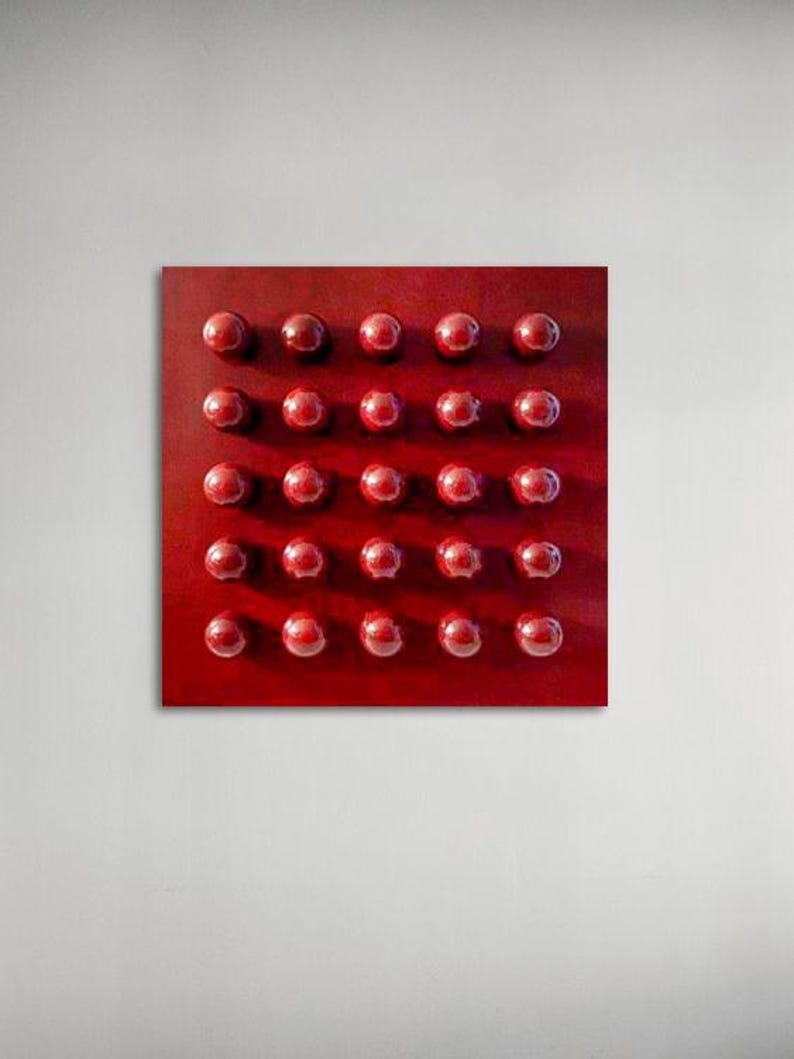 May include: A square, red abstract artwork with a grid of thirty-two raised, spherical shapes. The spheres are a darker shade of red than the background, creating a textured, dimensional effect. The artwork is mounted on a white wall.