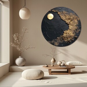 May include: A round black and gold abstract painting with a textured surface. The painting features a large, circular, gold shape in the upper right corner. The painting is hanging on a beige wall in a modern living room setting.