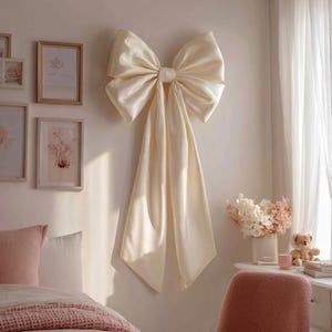 May include: A large, cream-colored satin bow is mounted on a wall. The bow has a voluminous design with long, flowing tails. The bow is the focal point of a bedroom, with framed artwork and a bed visible in the background.