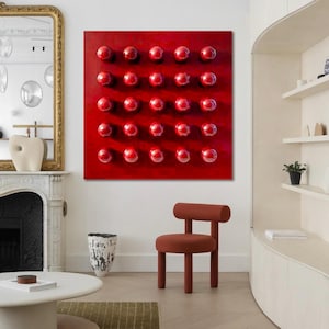 May include: A contemporary interior with a striking red artwork adorned with numerous raised spheres. A small, terracotta-hued chair is positioned in front of the artwork. The room also features a white coffee table, a gold-framed mirror, and a decorative vase.