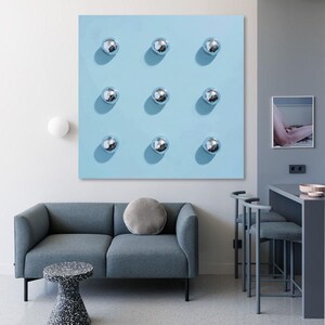 May include: A modern interior scene featuring a light blue wall art with nine silver disco balls arranged in a grid. A gray sofa with a round pillow, a speckled side table, and a bar area with stools complete the contemporary design.