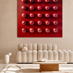 May include: A large, square, red abstract artwork featuring a grid of 30 raised, glossy, red spheres. The piece hangs above a beige sofa and a wooden coffee table in a modern living room setting.