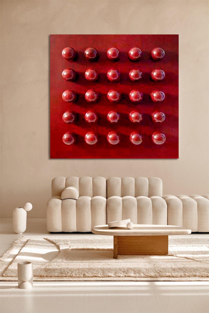May include: A large, square, red abstract artwork featuring a grid of 30 raised, glossy, red spheres. The piece hangs above a beige sofa and a wooden coffee table in a modern living room setting.