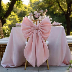 May include: A light pink chair bow decorates a gold chair at a wedding reception. The bow is made of soft fabric and is tied in a large bow shape. A light pink tablecloth and floral arrangement are also visible.