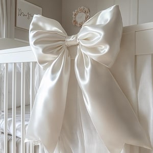 Könnte beinhalten: Eine große, elfenbeinfarbene Satinschleife ist an einem weißen Babybett befestigt. Die Schleife hat eine voluminöse Form mit langen, fließenden Bändern. Das Babybett ist weiß mit vertikalen Sprossen. Der Hintergrund umfasst einen gerahmten Druck und einen Blumenkranz.