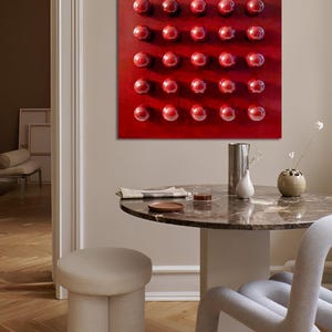 May include: A square, red abstract painting featuring a grid of 25 raised, glossy, red spheres. The artwork hangs above a round marble table with a beige stool and a grey chair. The room has a neutral colour palette.
