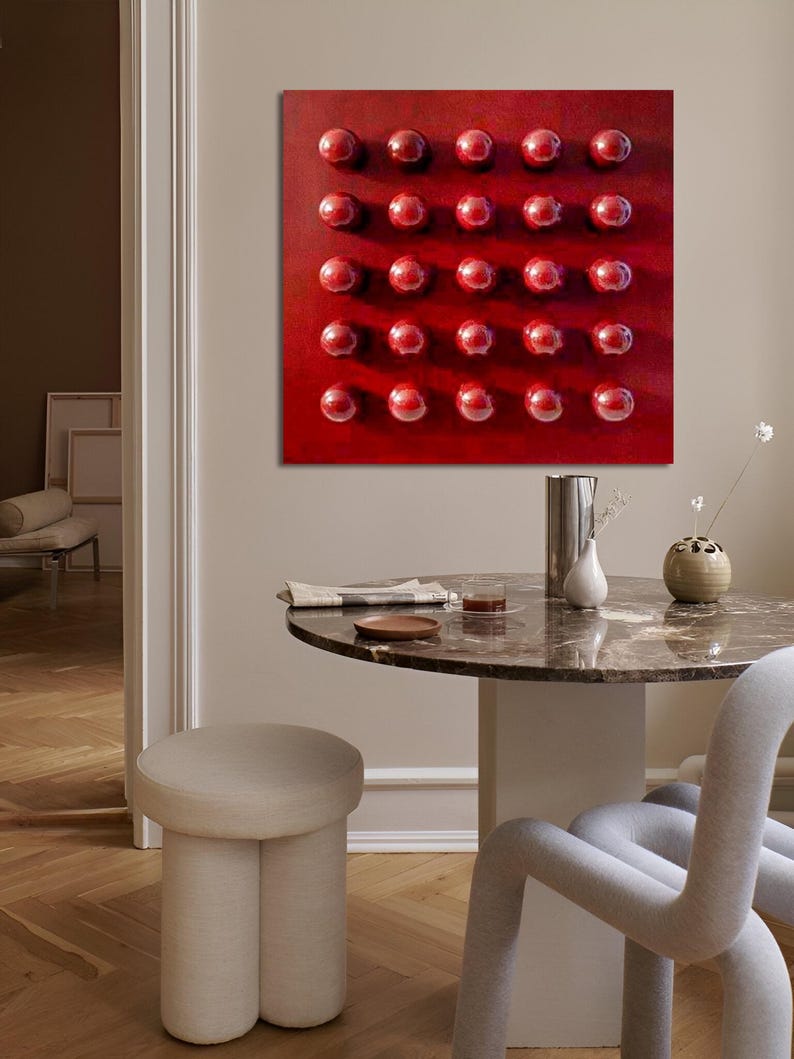 May include: A square, red abstract painting featuring a grid of 25 raised, glossy, red spheres. The artwork hangs above a round marble table with a beige stool and a grey chair. The room has a neutral colour palette.