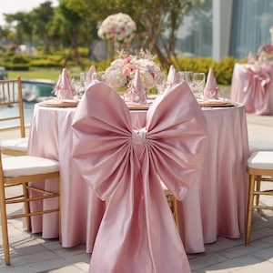 Puede incluir: Una mesa de recepción de bodas con una gran funda de silla con lazo rosa. La mesa está decorada con cargadores dorados, servilletas rosas y un centro de mesa floral. Las sillas tienen marcos dorados y asientos blancos.