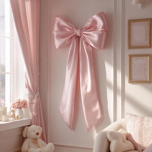 May include: A large, pale pink satin bow is mounted on a white wall. The bow has long, flowing tails. The room is decorated in soft pink tones, with matching curtains and a teddy bear visible in the background. Two framed pictures hang on the wall.