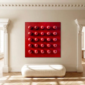 May include: A square, red abstract artwork featuring a grid of thirty-two raised, spherical, red elements. The piece is mounted on a light-coloured wall in a room with a white bench and hardwood flooring. The room has architectural details.