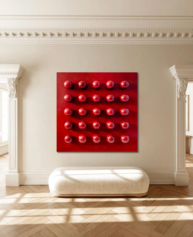 May include: A square, red abstract artwork featuring a grid of thirty-two raised, spherical, red elements. The piece is mounted on a light-coloured wall in a room with a white bench and hardwood flooring. The room has architectural details.