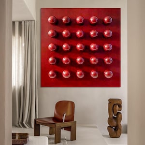 May include: A contemporary interior with a large, red square artwork featuring a grid of 30 raised, glossy red spheres. A brown leather chair and a wooden sculpture are in the foreground. The room has neutral walls and a light-coloured rug.
