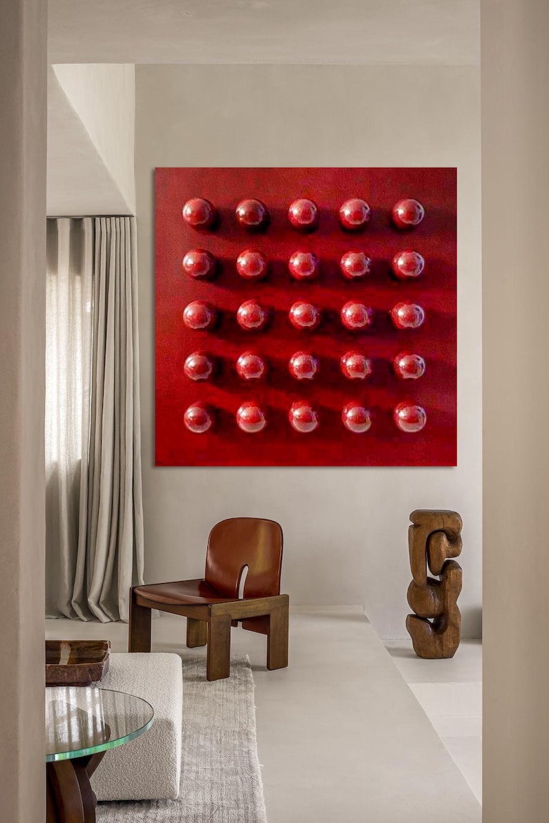 May include: A contemporary interior with a large, red square artwork featuring a grid of 30 raised, glossy red spheres. A brown leather chair and a wooden sculpture are in the foreground. The room has neutral walls and a light-coloured rug.