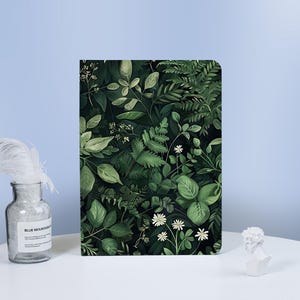以下が含まれることがあります： 緑豊かな植物のデザインが施された長方形のノート。表紙には、さまざまな葉、シダ、小さな白い花が描かれ、密度の高い自然なパターンを作り出しています。カラーパレットは、暗い背景に対して緑と白の色合いで構成されています。