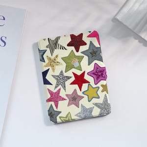 May include: A rectangular protective case for an e-reader featuring an off-white background covered in a pattern of multi-coloured stars. The stars appear in various textures and shades, including red, yellow, pink, silver, blue, and green.