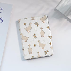 Puede incluir: Funda blanca para lector electrónico con un patrón de gansos y pequeñas bayas marrones.