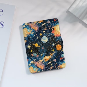 以下が含まれることがあります： カラフルな宇宙デザインのノート。表紙には、青、オレンジ、黄色の惑星、星、星雲が、濃い青色の背景に描かれています。ノートはマグネット開閉式です。