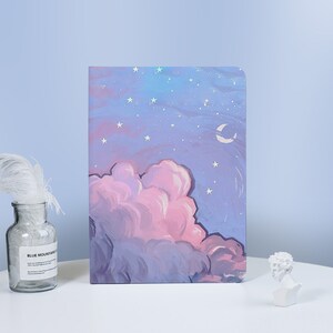 May include: A purple and pink notebook with a painted design of a night sky with clouds, stars, and a crescent moon.