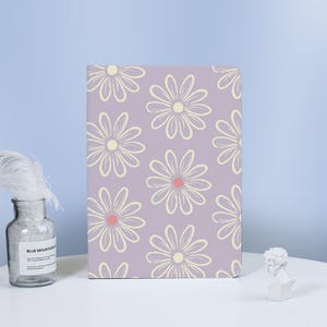 May include: A lavender notebook with a repeating pattern of cream-colored daisy flowers with pink centers. The notebook is standing upright on a white surface, with a glass bottle and a small white bust in the background.