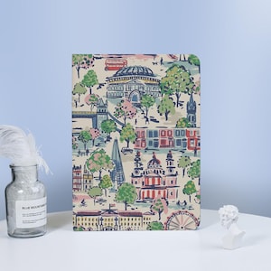 May include: A notebook featuring a colorful London-themed design. The cover showcases iconic landmarks like the London Eye, Tower Bridge, and red double-decker buses, set against a cream background with green and pink trees.