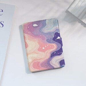 Könnte beinhalten: Ein geschlossenes, rechteckiges Etui mit einem farbenfrohen, wirbelnden Design. Das Design zeigt Farbtöne von Rosa, Lila und Blau mit weißen Akzenten, die an Wolken und Sterne erinnern. Das Etui scheint aus einem glatten Material zu bestehen.