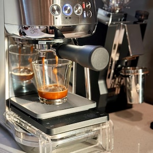 Puede incluir: Una máquina de espresso Breville plateada con una bandeja de goteo de acero inoxidable y un soporte de bandeja de goteo de plástico transparente. La máquina tiene una manija negra y un portafiltro negro y plateado. Dos tazas de vidrio transparentes están llenas de espresso.