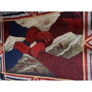 May include: A woven tapestry featuring a central graphic of two red hands gripping each other's wrists against a landscape background. The scene includes stylized mountains, a small house, and a deer silhouette, all framed by a decorative geometric border.