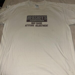 May include: Off-white t-shirt featuring the Hershey's Milk Chocolate logo with the text "MAY CAUSE ATTITUDE ADJUSTMENT" in black. The shirt is made of a soft, comfortable material.