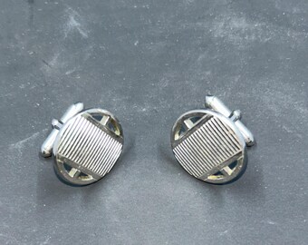 May include: A pair of silver cufflinks with a circular design. Each cufflink features a striped center and a geometric pattern around the edge. The cufflinks are displayed on a dark gray surface.