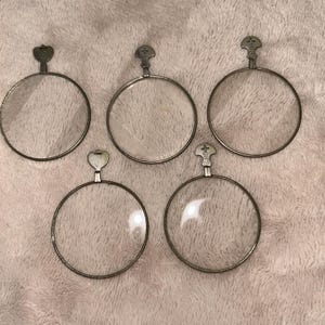 May include: Five vintage, circular eyeglasses lenses with metal frames. Each lens has a small handle at the top with a cross-shaped design. The lenses are clear, and the frames are a silver color. The items are arranged on a pinkish-beige surface.