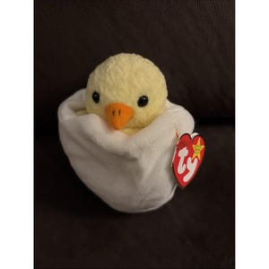 May include: A plush yellow chick with black eyes and an orange beak peeks out of a white eggshell. A red heart-shaped tag with the word "Ty" is attached to the egg. The stuffed animal is on a dark brown surface.