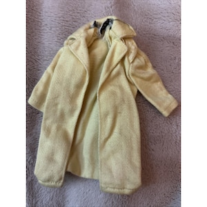 May include: A pale yellow coat with a collar and long sleeves. The coat is open in the front, revealing the interior lining. The fabric has a textured appearance. The coat is likely for a doll or small figure.