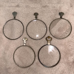 May include: Five antique eyeglasses with circular lenses and ornate metal frames. Each frame has a unique handle design, including heart, cross, and teardrop shapes. The lenses are clear, and the frames have a vintage, tarnished appearance.
