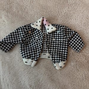 May include: A black and white houndstooth pattern jacket with a white collar featuring black polka dots and a pink accent. The jacket has silver buttons and is made of a knitted material.