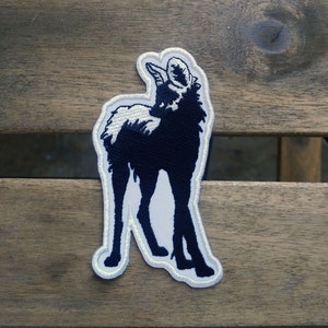 May include: Embroidered patch featuring a stylized ram in navy blue and white. The ram is depicted in profile, looking upwards, with a white outline and a light gray border. The patch is likely for clothing or accessories.