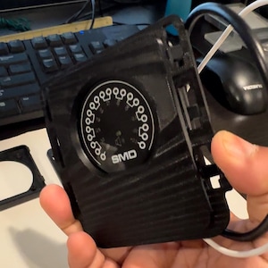 Puede incluir: Un dispositivo electrónico negro con un dial circular y las letras "SMD" impresas. El dial tiene marcas blancas y una aguja. El dispositivo se sostiene en la mano de una persona.