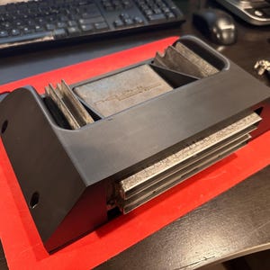 May include: A black electronic device with silver components, resting on a red cutting board. The device has a rectangular shape with a series of parallel metal fins. The background includes a keyboard, mouse, and other office items.