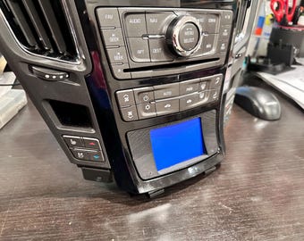 May include: A black car dashboard with a digital display screen, buttons for audio, navigation, climate control, and other functions. The screen is blue and shows a menu.