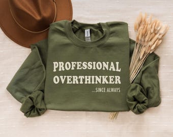 May include: An olive green sweatshirt with the text "PROFESSIONAL OVERTHINKER ...SINCE ALWAYS" in white. A brown hat and dried wheat stalks are also in the image. The sweatshirt is folded on a cream-colored surface.