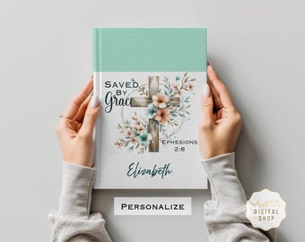 May include: A personalised journal cover featuring a watercolour illustration of a wooden cross adorned with flowers and the text "Saved by Grace Ephesians 2:8". The cover is light blue and the text is in black.